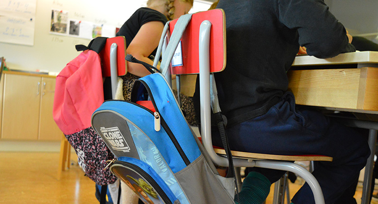 Två elever vid sina skolbänkar. Ryggsäckar hänger på stolarna. Foto.