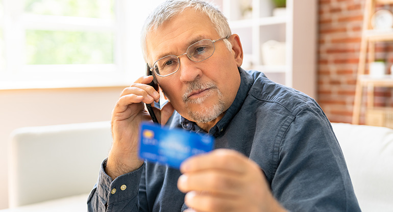 En äldre man håller i ett bankkort och pratar i telefon. Foto.